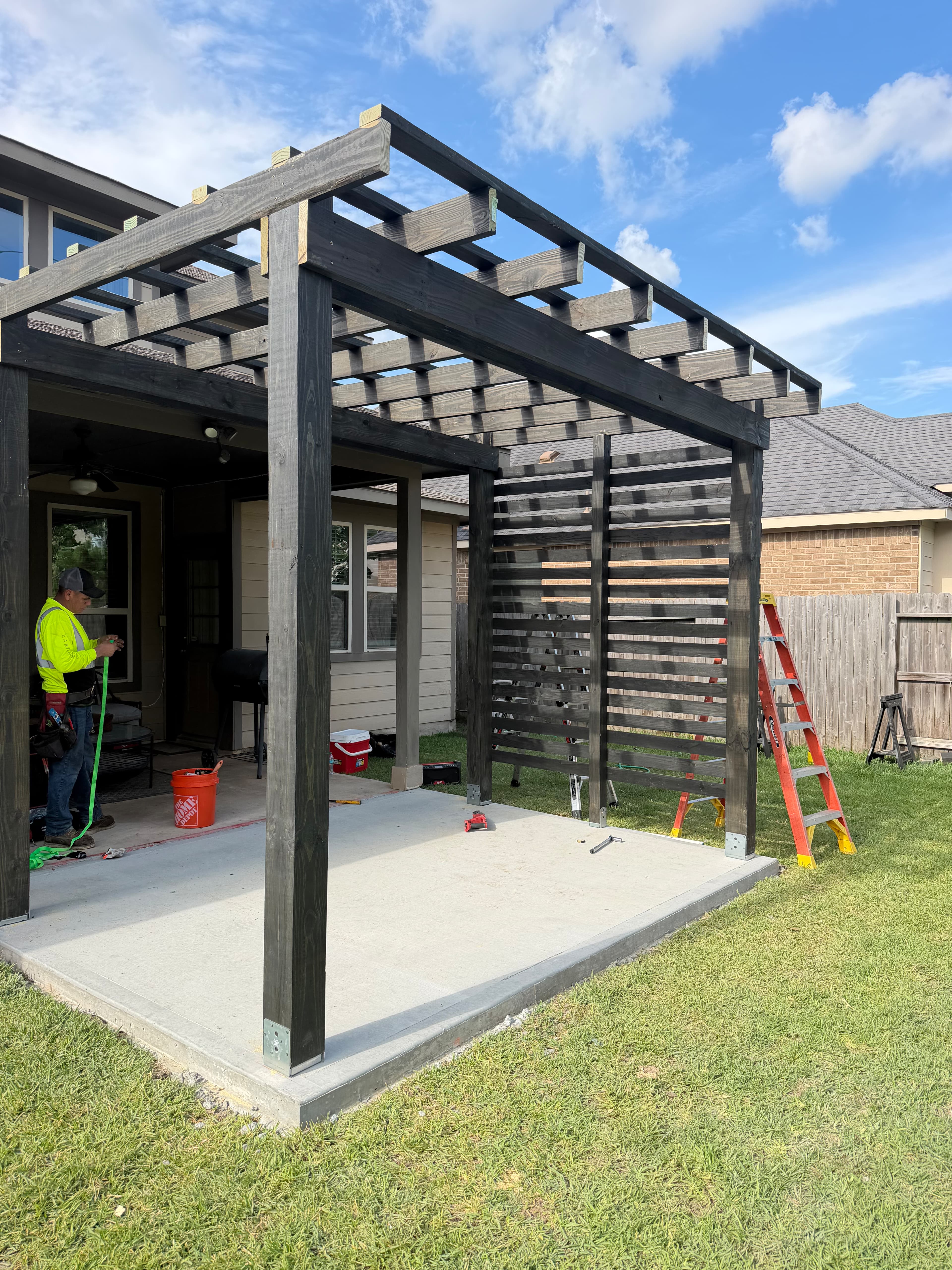 Dark-stained wood pergola under construction on a concrete patio with an orange ladder.