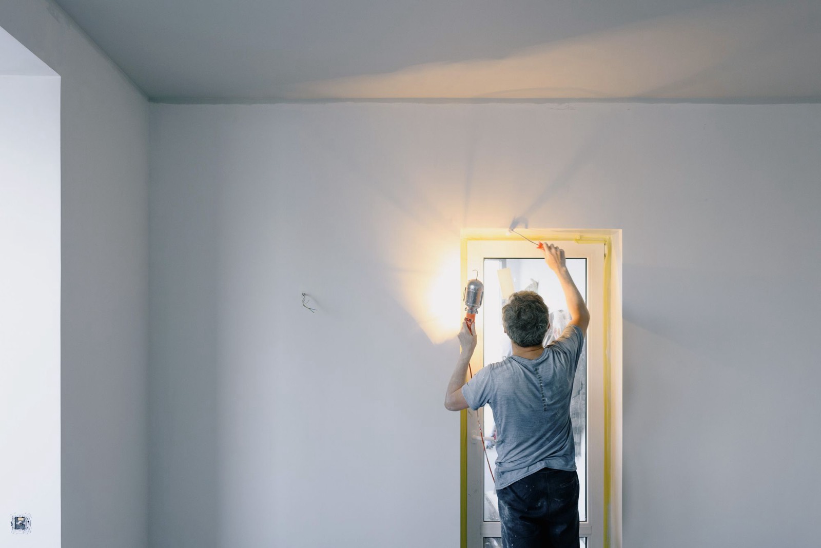 A person with their back to the camera working on a taped-off door frame in an empty room, lit by a handheld work light